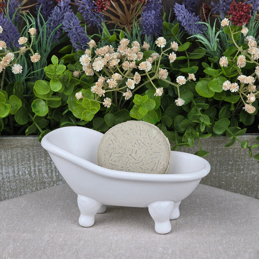 The Tea Tree Handmade Natural Shampoo & Conditioner Bar by Jarhead Aromatics, a round beige bar, is displayed in a small white ceramic bathtub with artificial green leaves and purple and white flowers in the background.