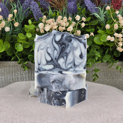Jarhead Aromatics’ Tea Tree + Tallow Handmade Artisan Soap bars, featuring detailed black, white, and gray marbling, are stacked on beige fabric with green plants and purple-cream flowers in the background for a natural look.