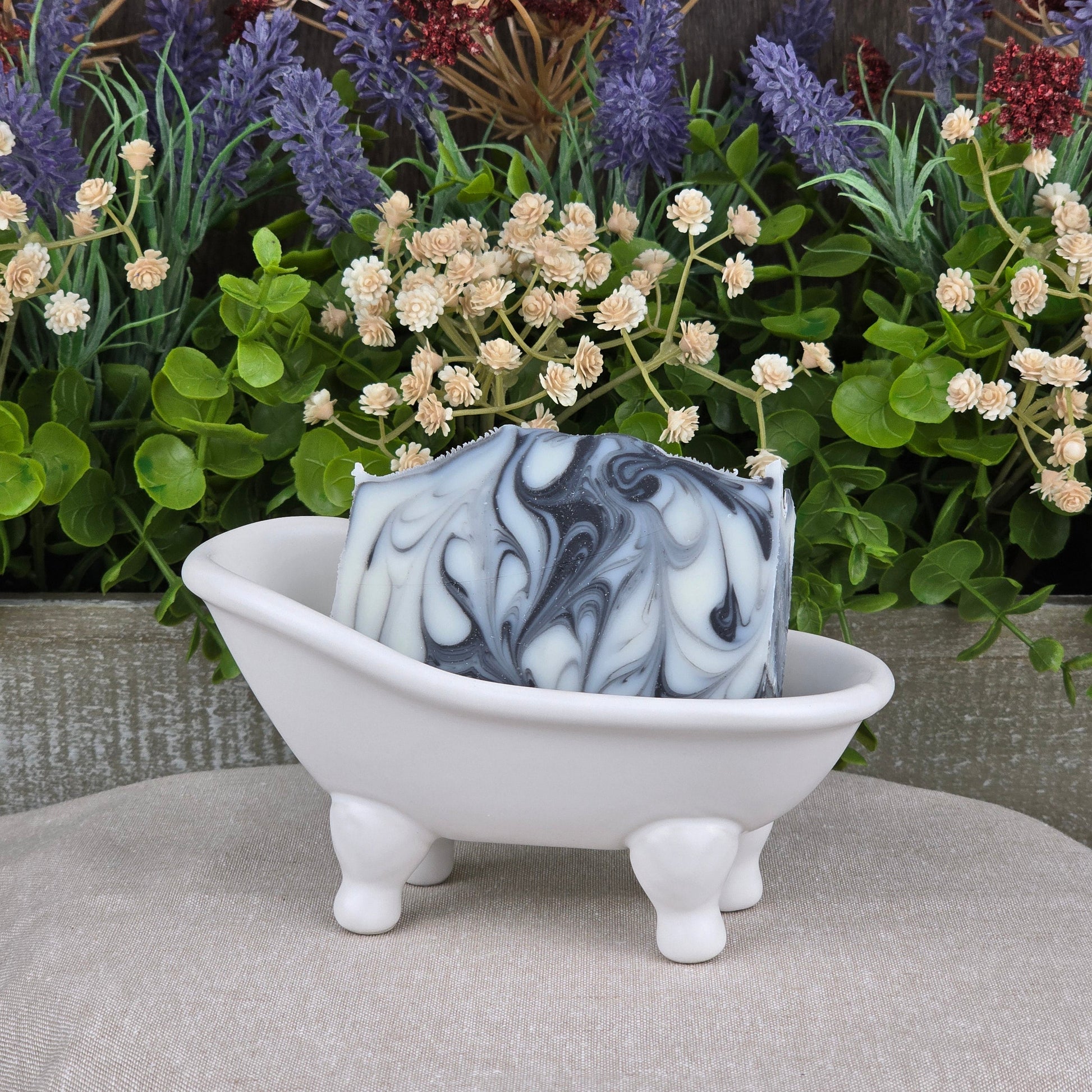 The Tea Tree + Tallow Handmade Artisan Soap by Jarhead Aromatics, featuring black and white marbling, rests in a small white bathtub-shaped dish with four legs, surrounded by green plants and purple and white artificial flowers.