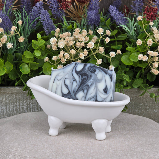 The Tea Tree + Tallow Handmade Artisan Soap by Jarhead Aromatics, featuring black and white marbling, rests in a small white bathtub-shaped dish with four legs, surrounded by green plants and purple and white artificial flowers.
