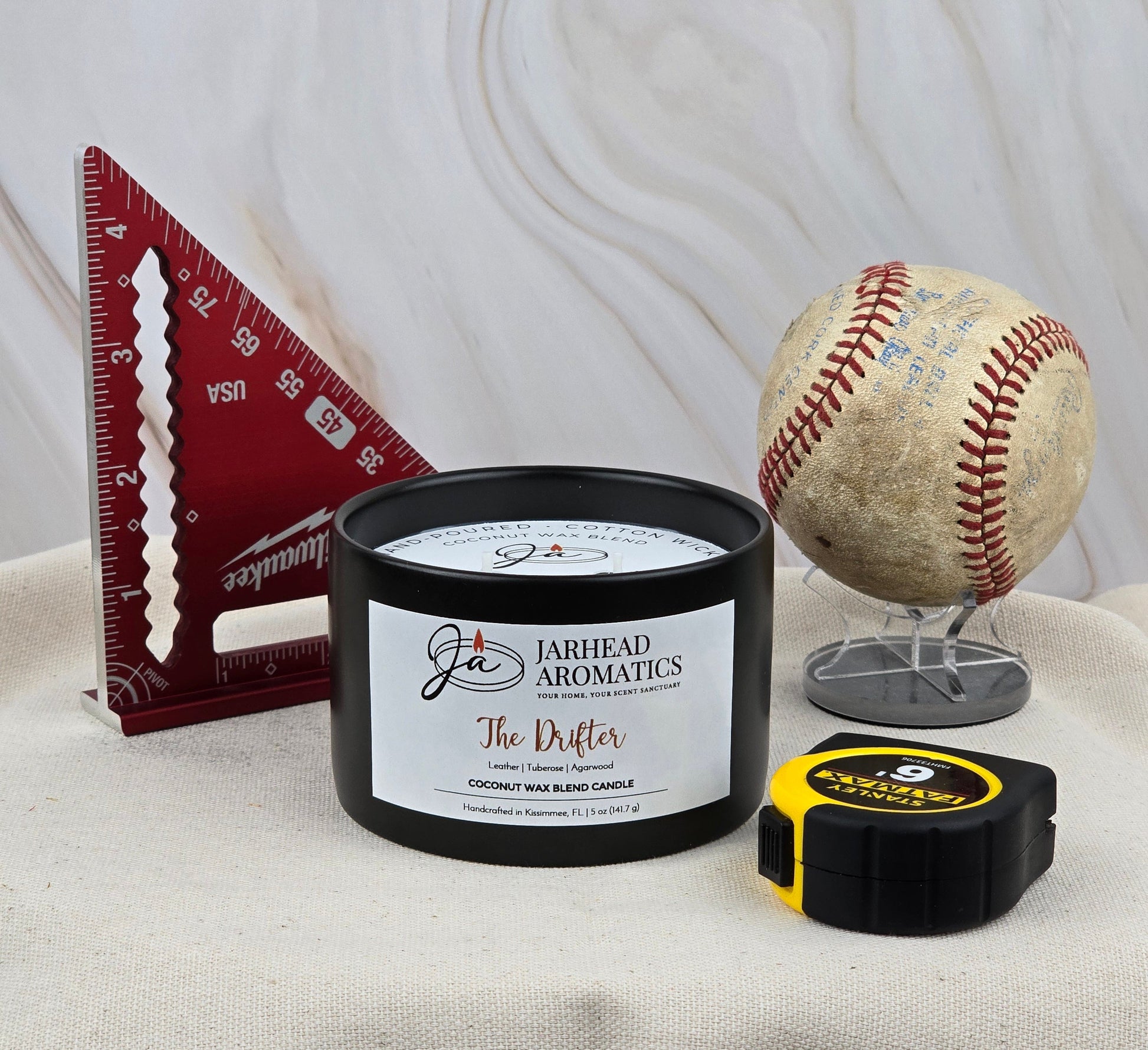 A baseball and a measuring tape evoke summer nostalgia, enhanced by the rugged Old West scent of The Drifter Handmade Non-Toxic Coconut Wax Blend Candle (5 oz ceramic) from Jarhead Aromatics.