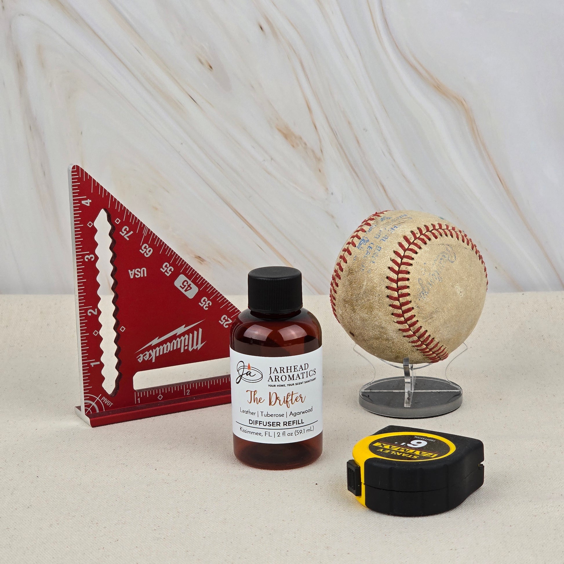 A red triangular ruler, a small bottle labeled “Jarhead Aromatics The Drifter Diffuser Refill | Leather, Tuberose & Agarwood,” an old baseball on a stand, and a black and yellow tape measure rest on a light surface with a marbled background.