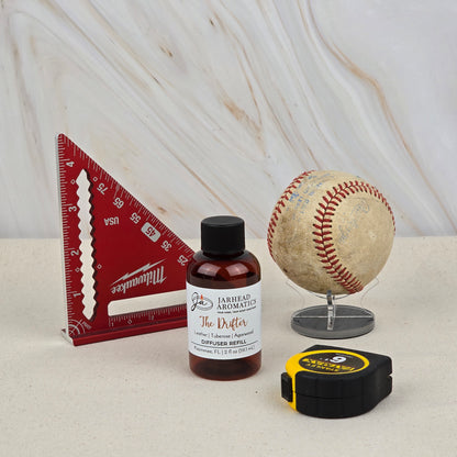 A red triangular ruler, a small bottle labeled “Jarhead Aromatics The Drifter Diffuser Refill | Leather, Tuberose & Agarwood,” an old baseball on a stand, and a black and yellow tape measure rest on a light surface with a marbled background.