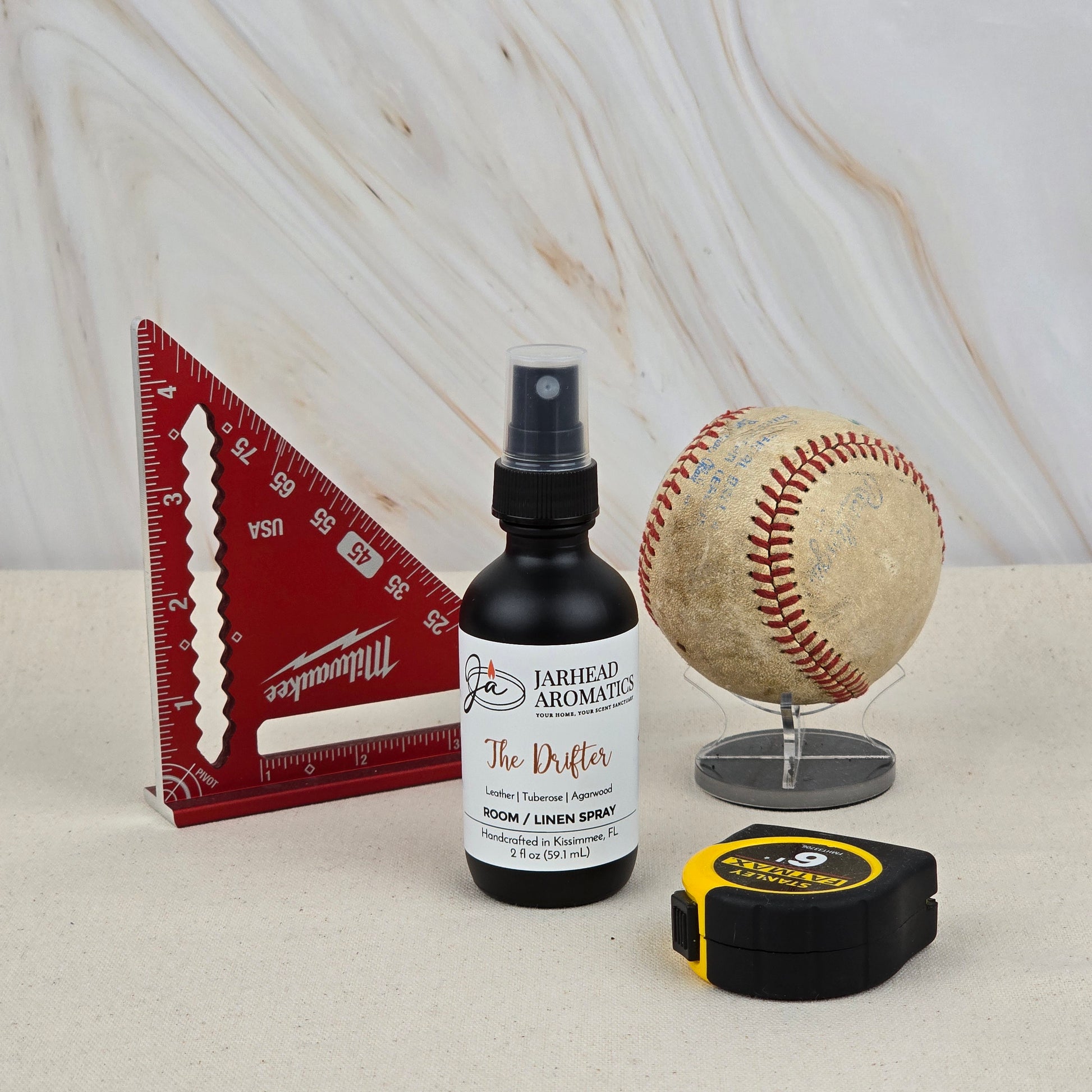 A bottle of The Drifter Room & Linen Spray by Jarhead Aromatics sits on a marble-style surface with a red triangular ruler, worn baseball on a stand, and a yellow-black tape measure.