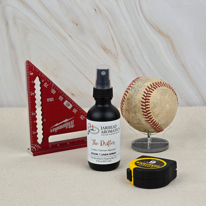 A bottle of The Drifter Room & Linen Spray by Jarhead Aromatics sits on a marble-style surface with a red triangular ruler, worn baseball on a stand, and a yellow-black tape measure.