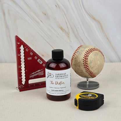 A red triangular ruler, a Jarhead Aromatics The Drifter Diffuser Refill (Leather, Tuberose & Agarwood), a baseball on a stand, and a black and yellow tape measure are displayed on a beige surface with a marbled background.