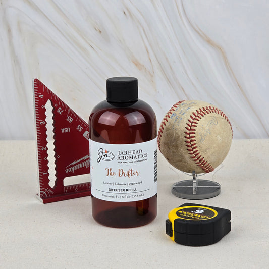 A bottle of The Drifter Diffuser Refill | Leather, Tuberose & Agarwood by Jarhead Aromatics sits beside a red triangular ruler, a baseball on a stand, and a yellow-and-black tape measure.