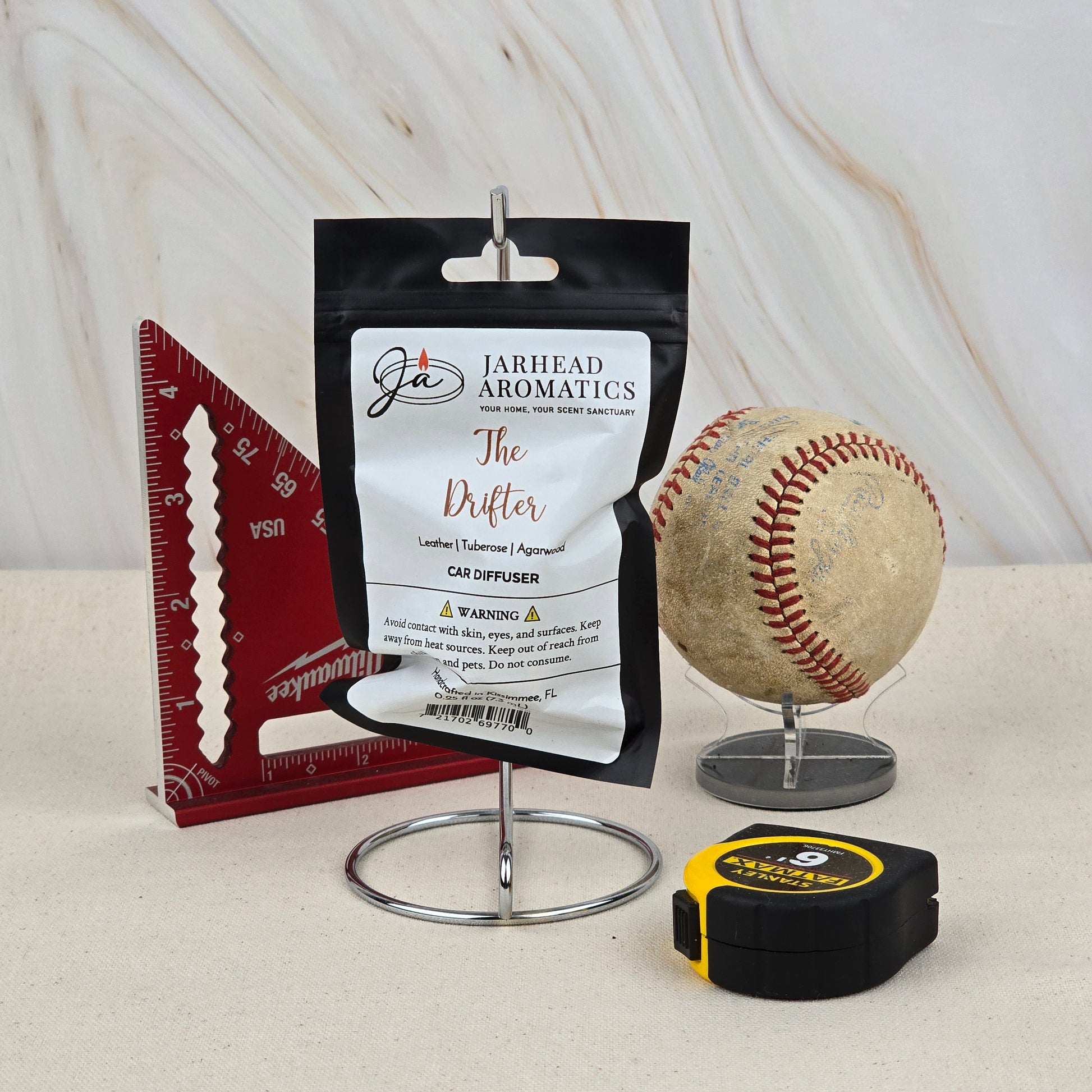 The Drifter Car Diffuser by Jarhead Aromatics, featuring leather, desert bloom & oud scent, hangs on a stand beside a red triangular ruler, a worn baseball in a clear holder, and a yellow-black tape measure on a light textured surface with marble background.