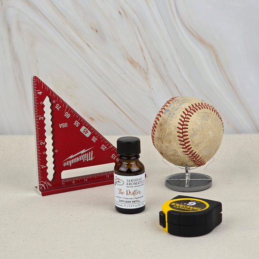 A red triangular ruler, a bottle of Jarhead Aromatics The Drifter Cold Air Diffuser Refill (Leather & Agarwood), an old baseball on a stand, and a yellow-black tape measure sit on a light surface with a marble-patterned background.