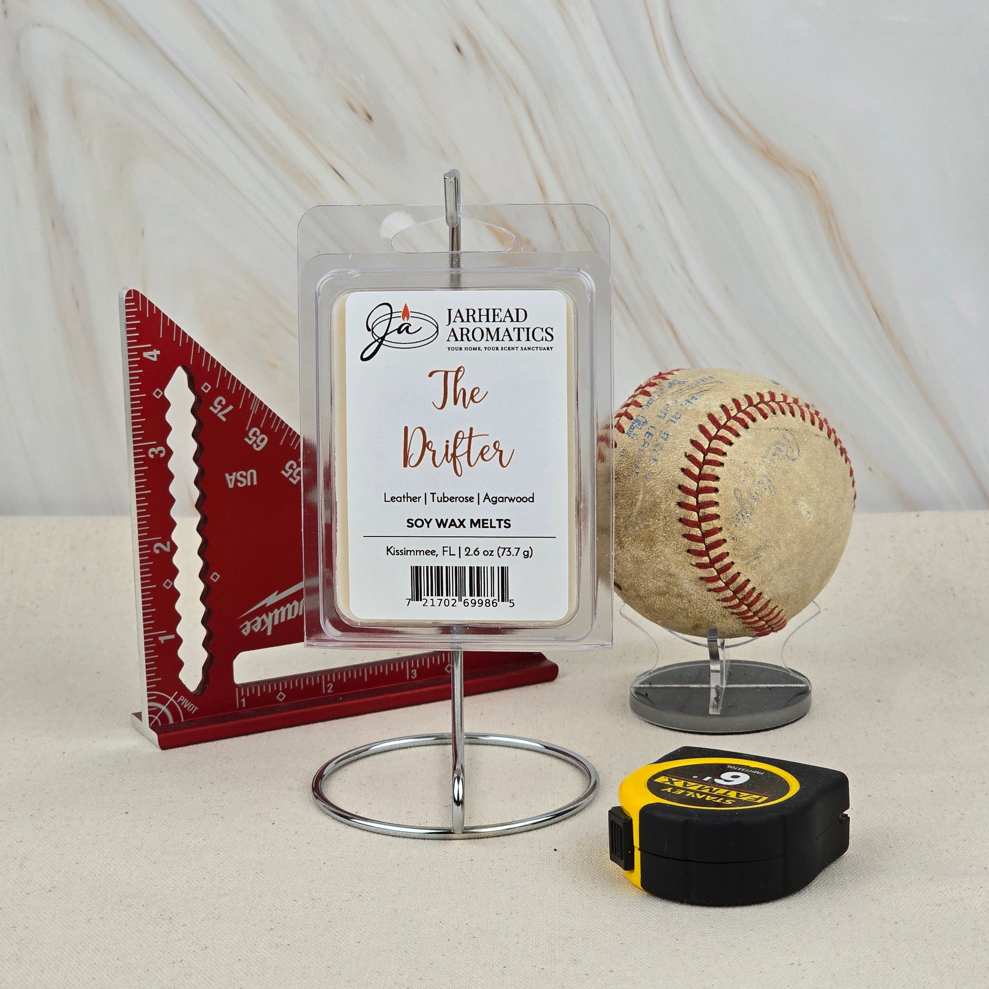 A Jarhead Aromatics The Drifter Soy Wax Melt | Leather & Agarwood Western Wax Melts is displayed on a stand beside a red triangular ruler, a worn baseball, and a yellow-black tape measure on a beige surface with a light backdrop.