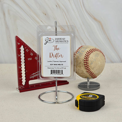 A Jarhead Aromatics The Drifter Soy Wax Melt | Leather & Agarwood Western Wax Melts is displayed on a stand beside a red triangular ruler, a worn baseball, and a yellow-black tape measure on a beige surface with a light backdrop.