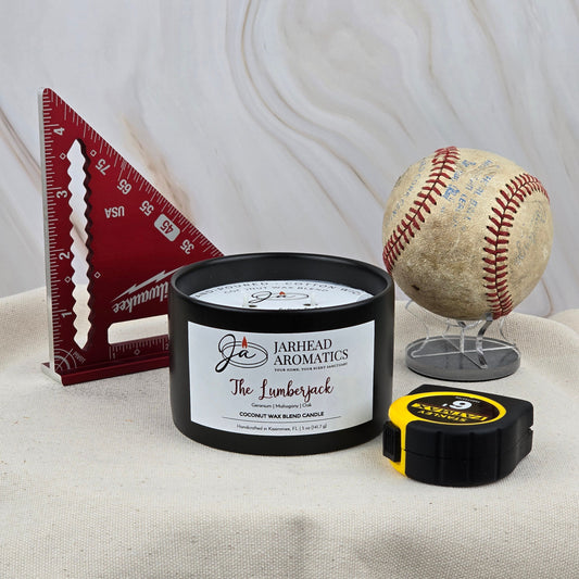 A red triangle ruler, Jarhead Aromatics The Lumberjack - Coconut Wax Blend Candle with mahogany scent, a yellow and black tape measure, and a worn baseball on a wire stand are arranged on beige fabric.
