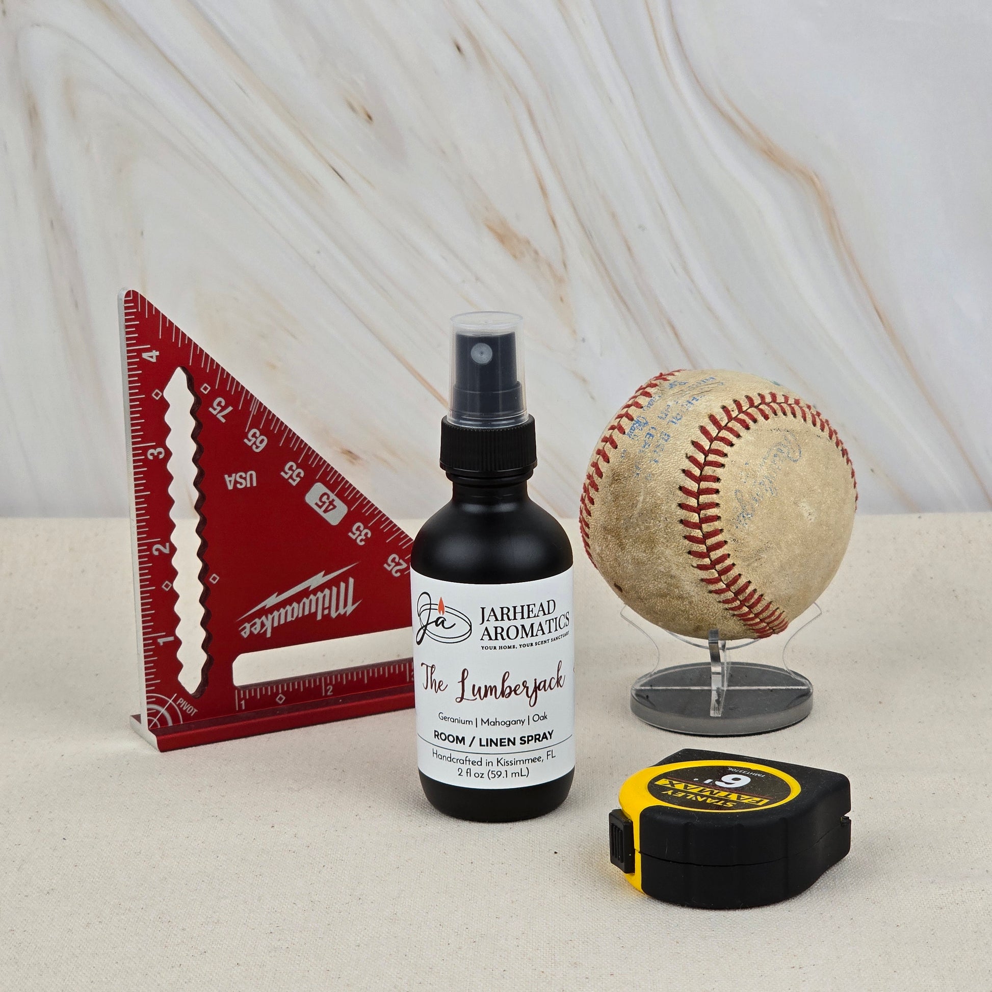 A red triangular ruler, a worn baseball on a stand, a black and yellow tape measure, and Jarhead Aromatics' The Lumberjack Room & Linen Spray with its woodsy mahogany, cedar & oak scent are arranged on a light surface against a marbled background.