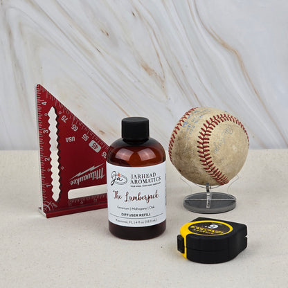 A red triangular ruler, Jarhead Aromatics' The Lumberjack Diffuser Refill with woodsy mahogany, cedar & oak scents, a baseball on a stand, and a black-and-yellow tape measure sit on a light surface.