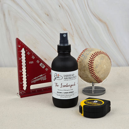 A black spray bottle labeled Jarhead Aromatics The Lumberjack Room & Linen Spray sits beside a red triangle ruler, an old baseball on a stand, and a yellow-and-black tape measure—perfect for this woodsy mahogany, cedar & oak fragrance.