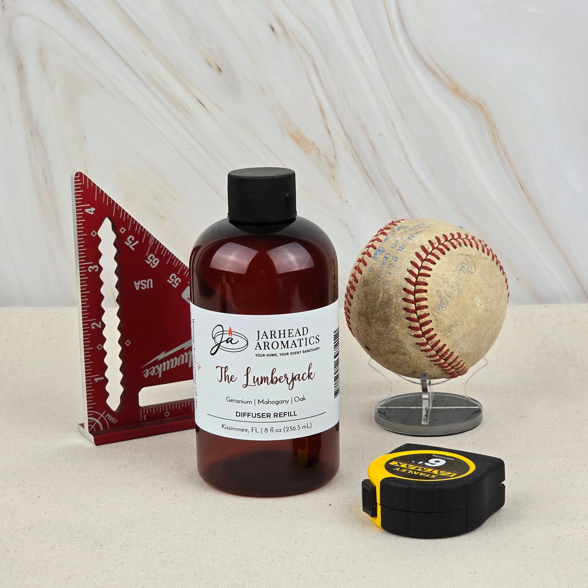 A bottle of Jarhead Aromatics The Lumberjack Diffuser Refill | Woodsy Mahogany, Cedar & Oak Home Fragrance Oil Refill sits on a beige surface with a red triangle ruler, baseball on a stand, and tape measure—evoking its woodland scent.
