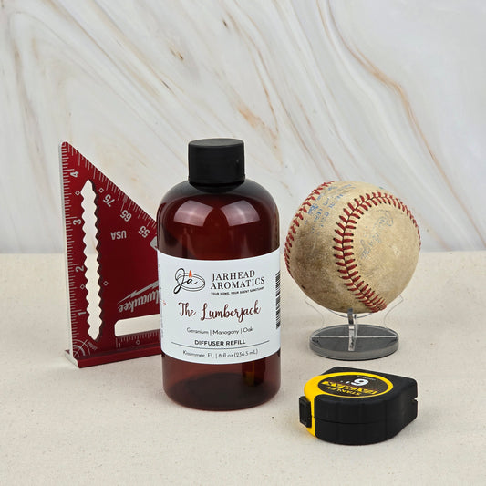 A bottle of Jarhead Aromatics The Lumberjack Diffuser Refill | Woodsy Mahogany, Cedar & Oak Home Fragrance Oil Refill sits on a beige surface with a red triangle ruler, baseball on a stand, and tape measure—evoking its woodland scent.
