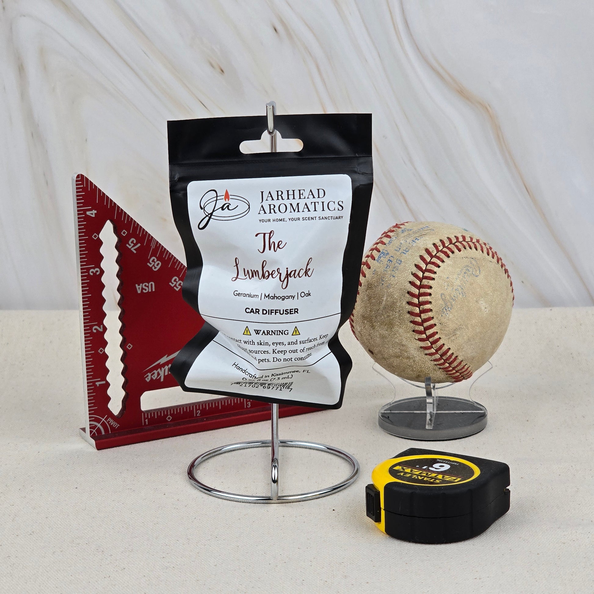 The Lumberjack Car Diffuser by Jarhead Aromatics, featuring cedar, oak & mahogany scents, rests on a metal holder beside a red triangular ruler, worn baseball on a stand, and yellow-black tape measure against a neutral backdrop.