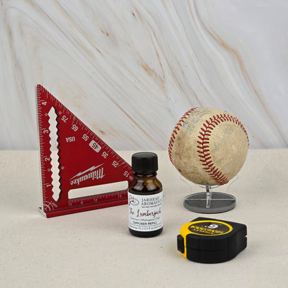 A red triangular ruler, a small bottle of Jarhead Aromatics The Lumberjack Mahogany & Cedarwood diffuser oil refill, an old baseball on a stand, and a black and yellow tape measure are arranged on a beige surface with a marbled background.