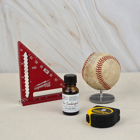 A red triangular ruler, a small bottle of Jarhead Aromatics The Lumberjack Mahogany & Cedarwood diffuser oil refill, an old baseball on a stand, and a black and yellow tape measure are arranged on a beige surface with a marbled background.