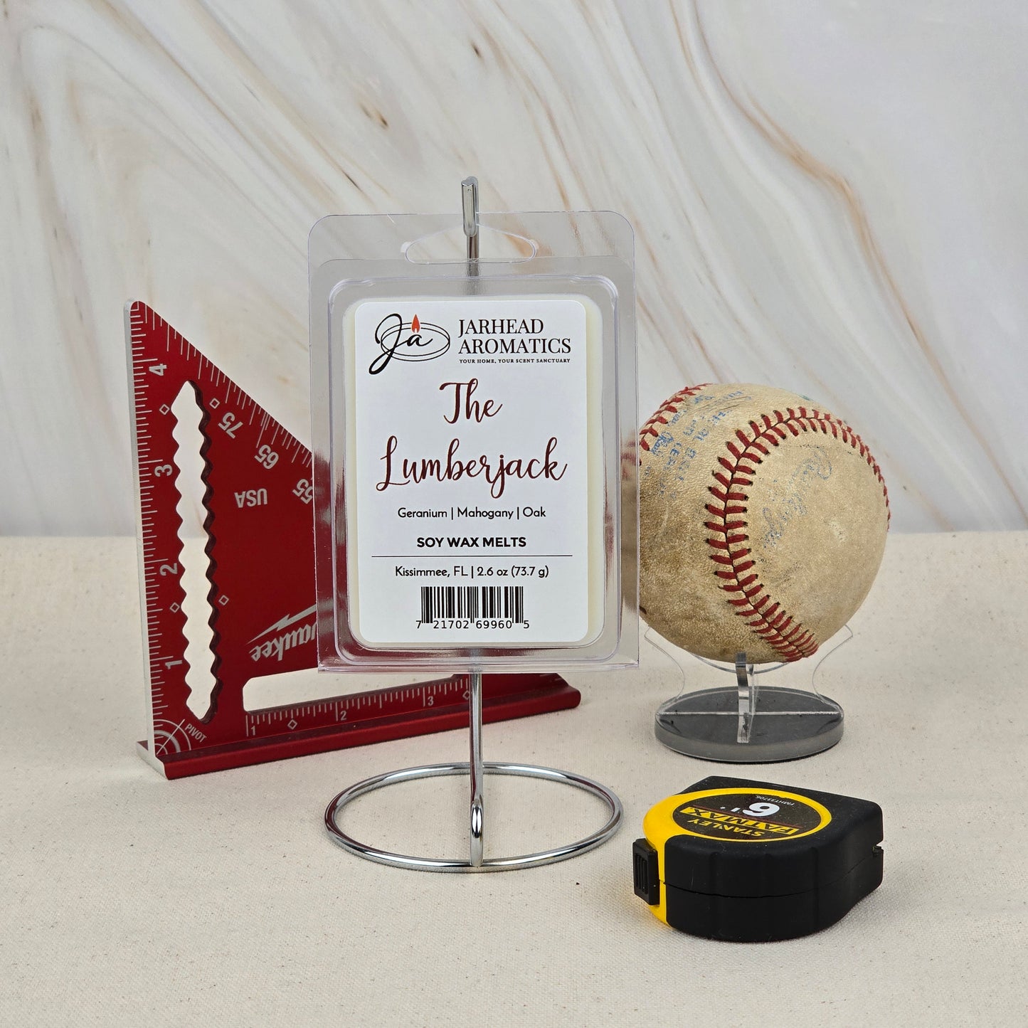 A package of Jarhead Aromatics The Lumberjack Soy Wax Melt | Mahogany Cedar & Oak is shown on a stand with a red ruler, worn baseball, and tape measure—emphasizing its hand-poured soy wax and rugged woodsy scent.