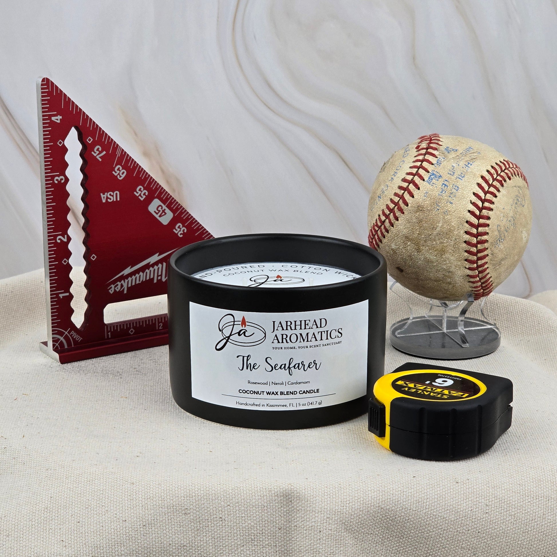 A red triangular ruler, a worn baseball on a stand, and Jarhead Aromatics' The Seafarer Handmade Non-Toxic Coconut Wax Blend Candle in a 5 oz ceramic container are arranged beside a yellow tape measure on a light surface with a marble background.