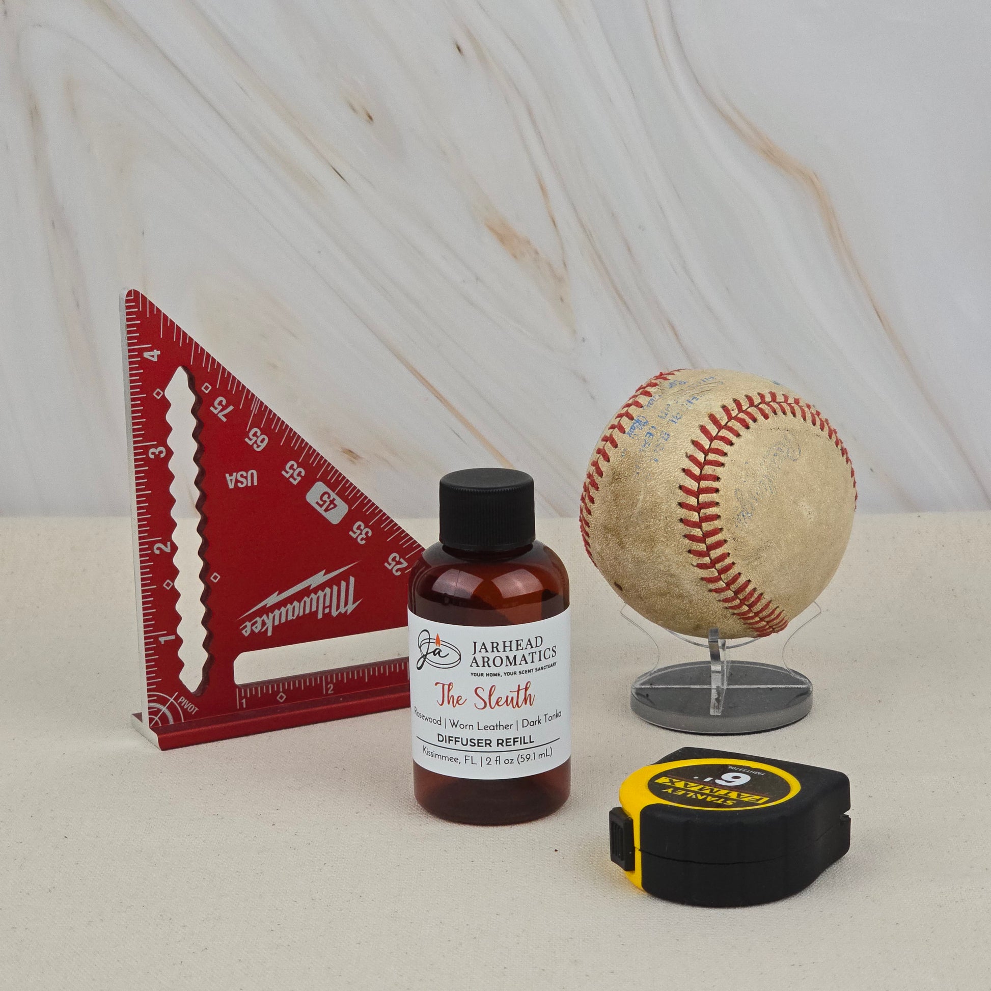 A red triangular ruler, The Sleuth Diffuser Refill by Jarhead Aromatics (Rosewood, Leather & Sandalwood fragrance), a baseball on a clear stand, and a yellow-black tape measure are arranged on a white surface with a marble background.