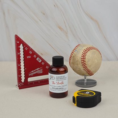 A red triangular ruler, The Sleuth Diffuser Refill by Jarhead Aromatics (Rosewood, Leather & Sandalwood fragrance), a baseball on a clear stand, and a yellow-black tape measure are arranged on a white surface with a marble background.