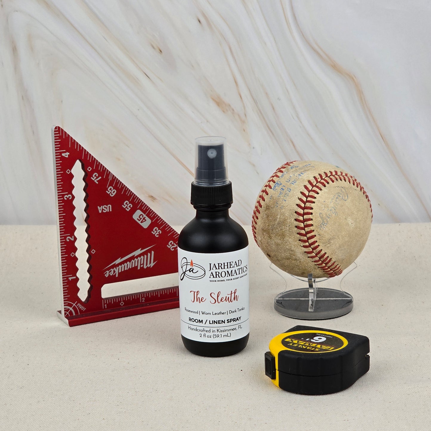 A red triangular ruler, a worn baseball on a stand, a black-and-yellow tape measure, and Jarhead Aromatics' The Sleuth Room & Linen Spray (Leather, Cedar & Sandalwood) are displayed on a light surface with a marble-patterned background.