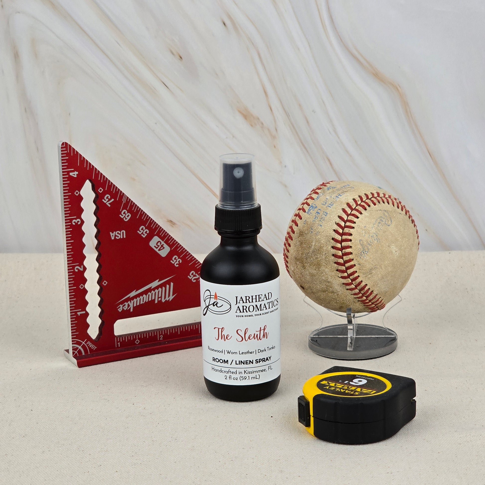 A red triangular ruler, a worn baseball on a stand, a black-and-yellow tape measure, and Jarhead Aromatics' The Sleuth Room & Linen Spray (Leather, Cedar & Sandalwood) are displayed on a light surface with a marble-patterned background.