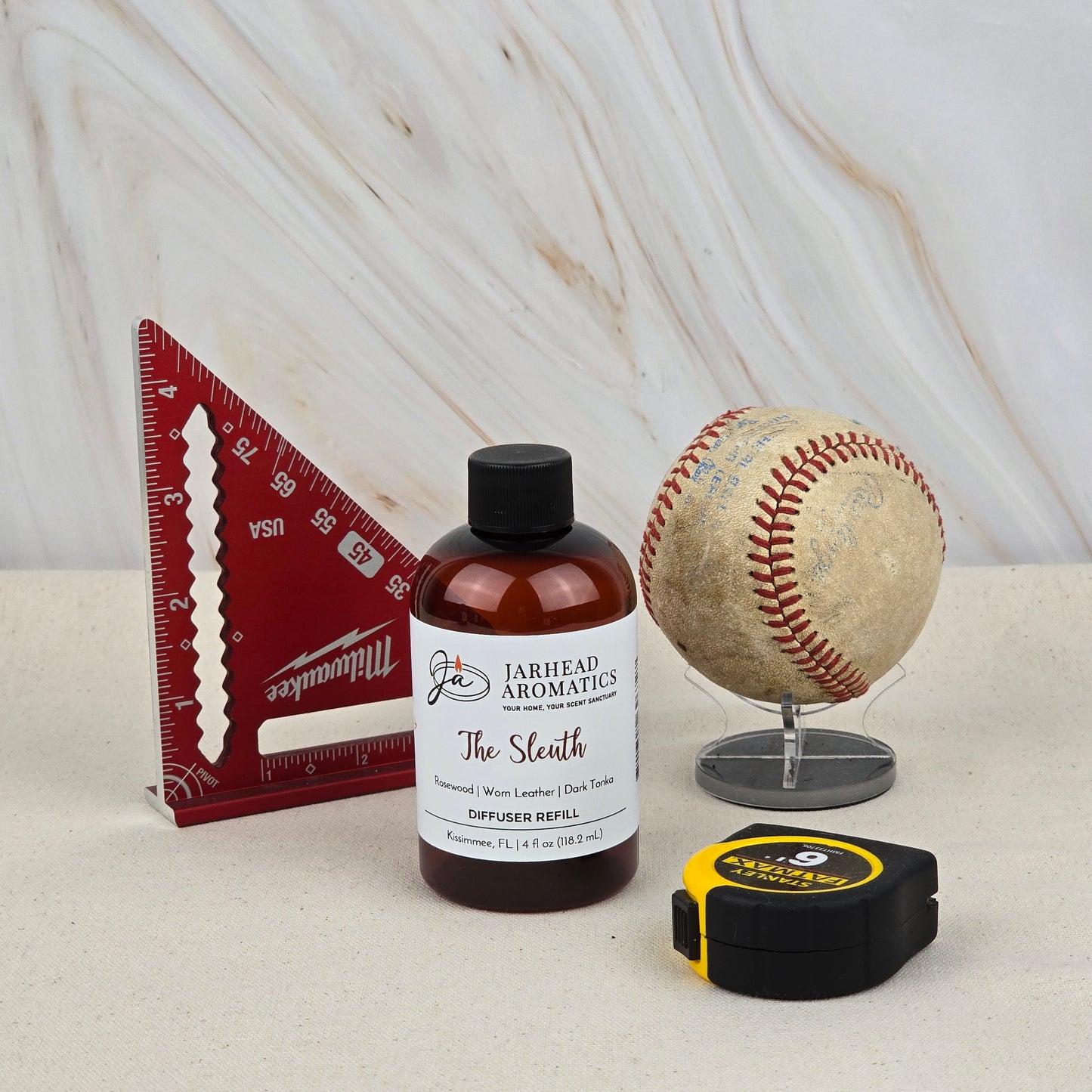 A red triangular ruler, a Jarhead Aromatics The Sleuth Diffuser Refill (Rosewood, Leather & Sandalwood), a baseball on a stand, and a black and yellow tape measure are displayed on a light surface with a marbled background.