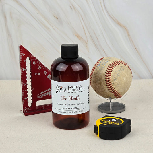 A bottle of Jarhead Aromatics The Sleuth Diffuser Refill | Rosewood, Leather & Sandalwood Mystery Home Fragrance Oil stands on a light surface beside a red triangular ruler, baseball on a stand, and black and yellow tape measure.