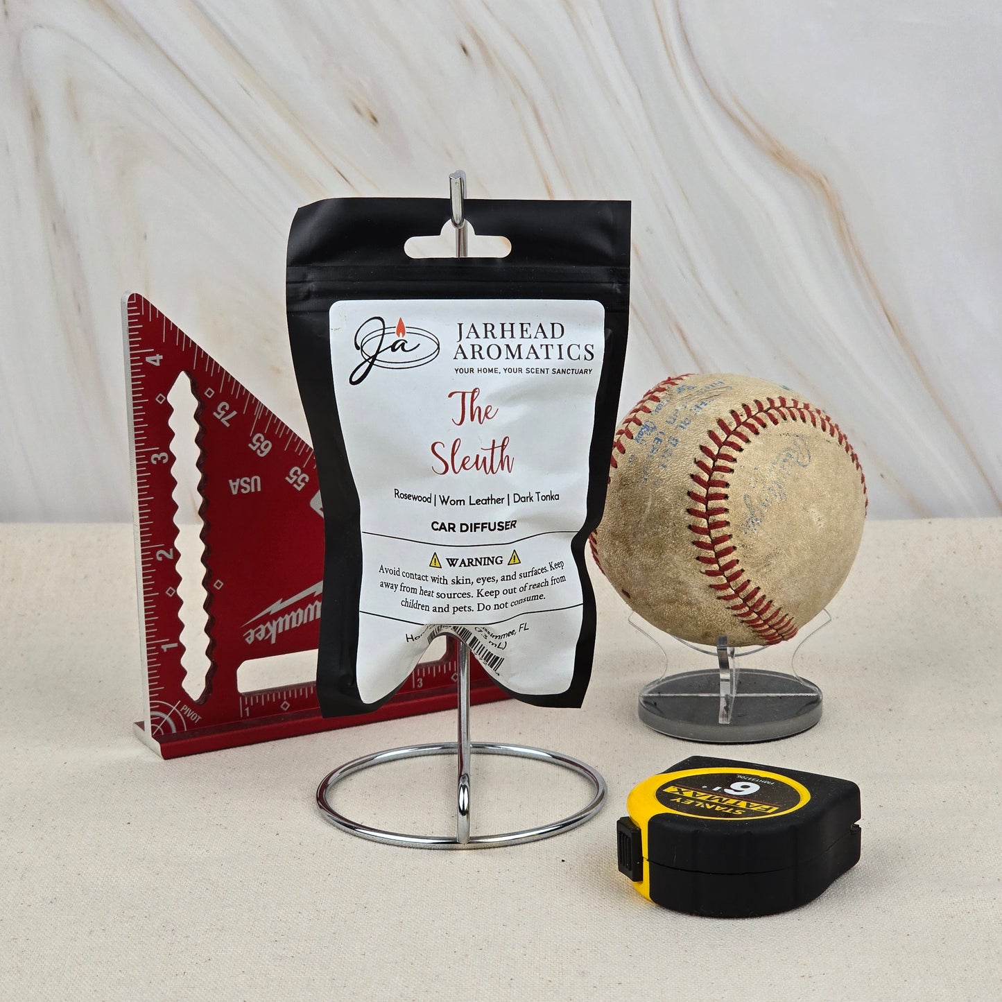 A Jarhead Aromatics The Sleuth Car Diffuser, featuring warm leather, cedar & sandalwood scents, stands on a metal holder beside a red triangular ruler, an old baseball on a clear stand, and a black and yellow tape measure.