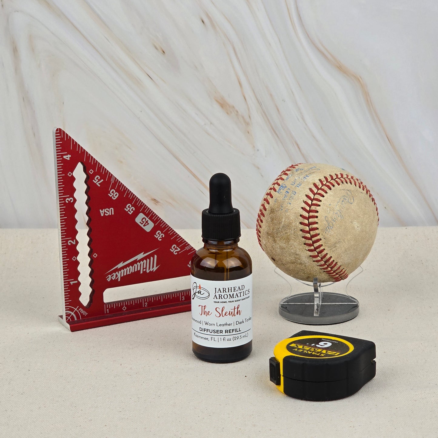 A red triangular ruler, Jarhead Aromatics' The Sleuth Car Diffuser Refill (Leather, Sandalwood & Patchouli), a worn baseball on a stand, and a yellow tape measure rest on a light surface with a marbled background.