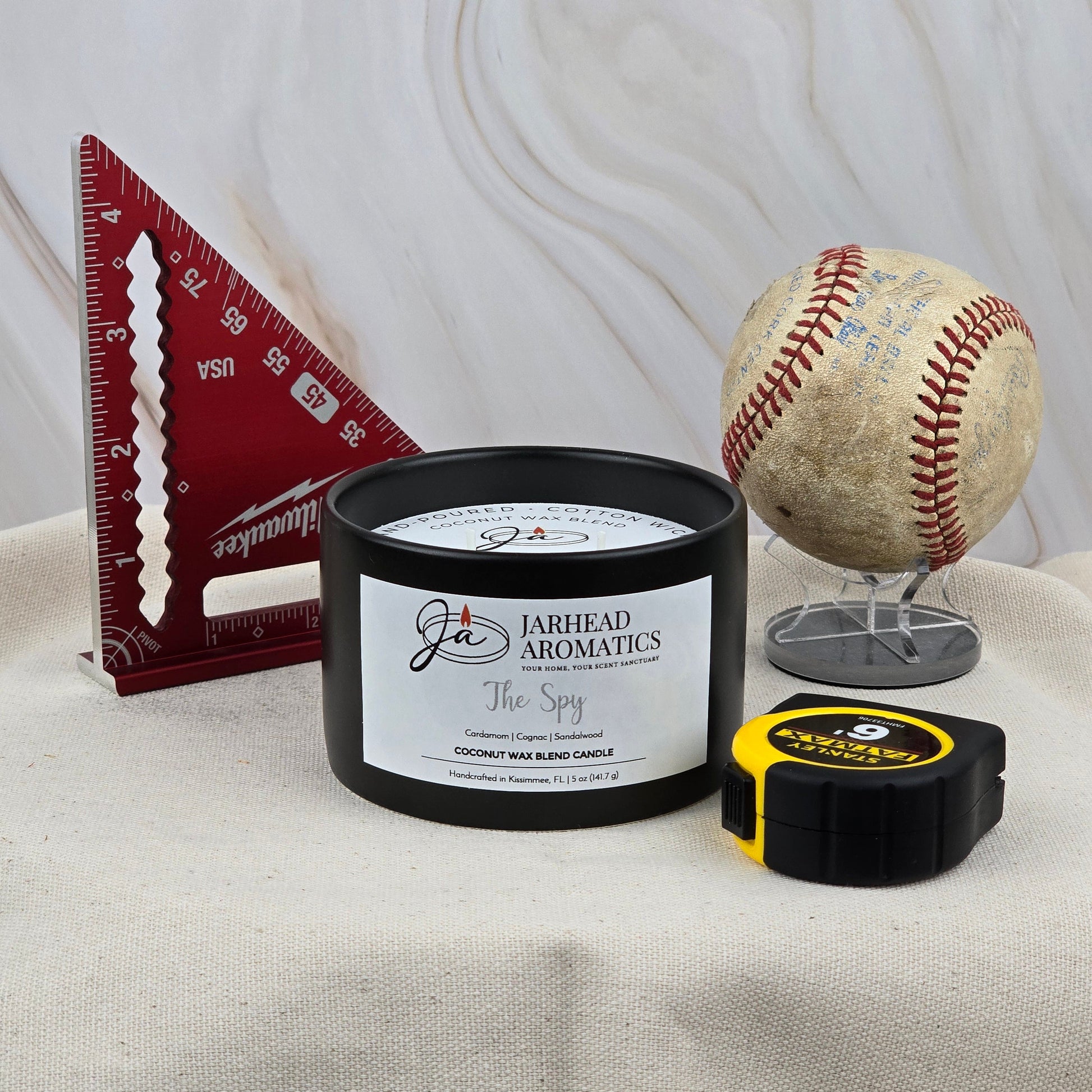 A 5 oz ceramic candle from Jarhead Aromatics labeled "The Spy" rests on fabric with a red measuring square, an old baseball, and a yellow tape measure—evoking the scent of leather and cedar.