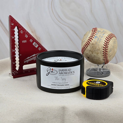 A 5 oz ceramic candle from Jarhead Aromatics labeled "The Spy" rests on fabric with a red measuring square, an old baseball, and a yellow tape measure—evoking the scent of leather and cedar.