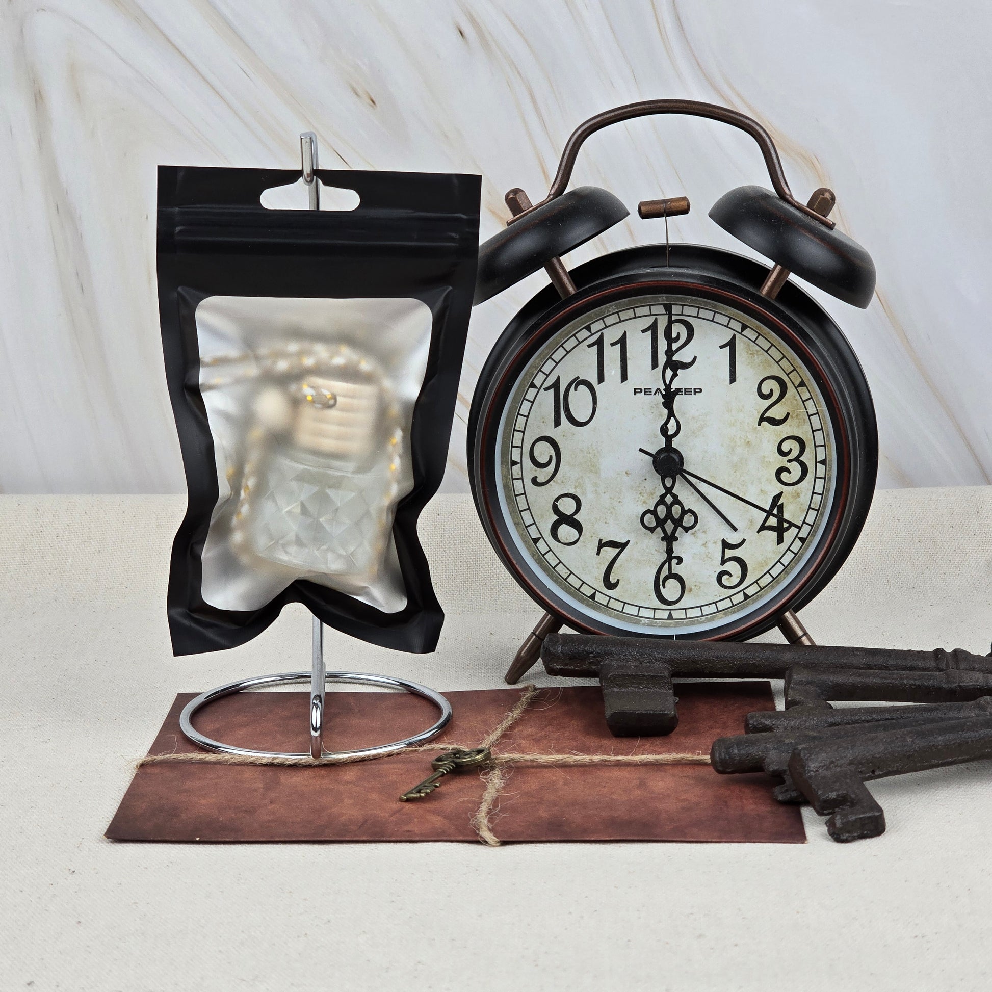 A black alarm clock, antique keys, a small key on an envelope, and a clear pouch holding Jarhead Aromatics’ Sweet Sunrise Car Diffuser | Bright Citrus & Peach Car Air Freshener are arranged on a light surface against a marble background.