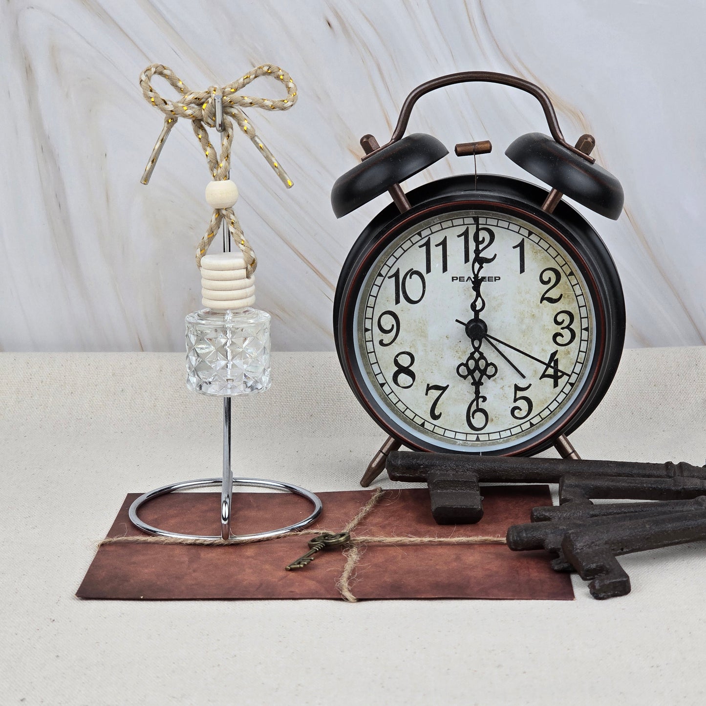 A vintage black alarm clock sits on a brown envelope with twine, beside Jarhead Aromatics’ Sweet Sunrise Car Diffuser (Bright Citrus & Peach) with a knotted rope and wooden beads, and two large decorative metal keys.