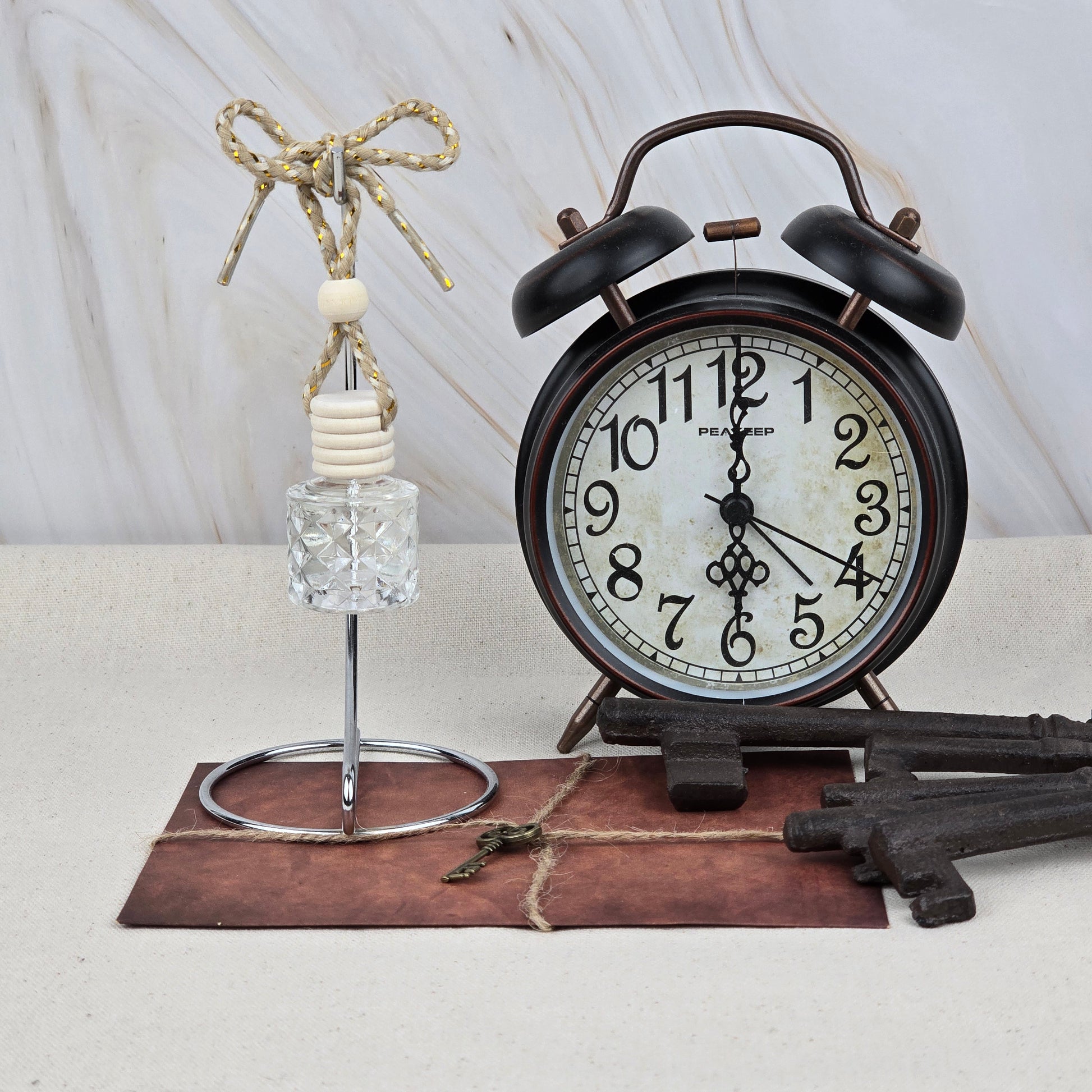 A vintage black alarm clock sits on a brown envelope with twine, beside Jarhead Aromatics’ Sweet Sunrise Car Diffuser (Bright Citrus & Peach) with a knotted rope and wooden beads, and two large decorative metal keys.