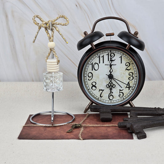 A vintage black alarm clock sits on a brown envelope with twine, beside Jarhead Aromatics’ Sweet Sunrise Car Diffuser (Bright Citrus & Peach) with a knotted rope and wooden beads, and two large decorative metal keys.