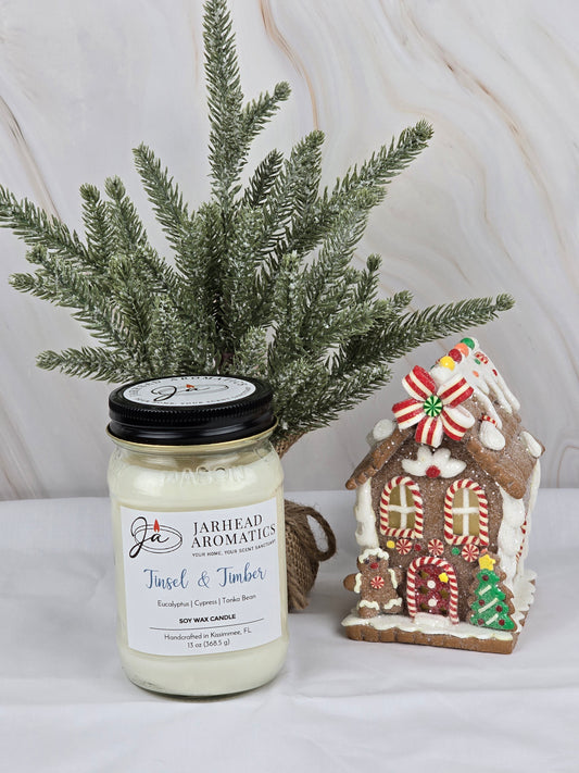 A Tinsel & Timber - Soy Wax Candle by Jarhead Aromatics, exuding eucalyptus, cypress, tonka bean, and a hint of mint, sits next to a gingerbread house and an evergreen branch, capturing the essence of holiday greenery.