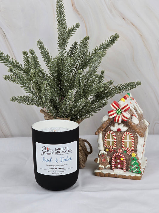 A Tinsel & Timber soy wax candle by Jarhead Aromatics in black sits beside a candy-adorned gingerbread house. A frosted pine tree fills the air with Christmas tree scent, creating a festive ambiance against the light marbled background.