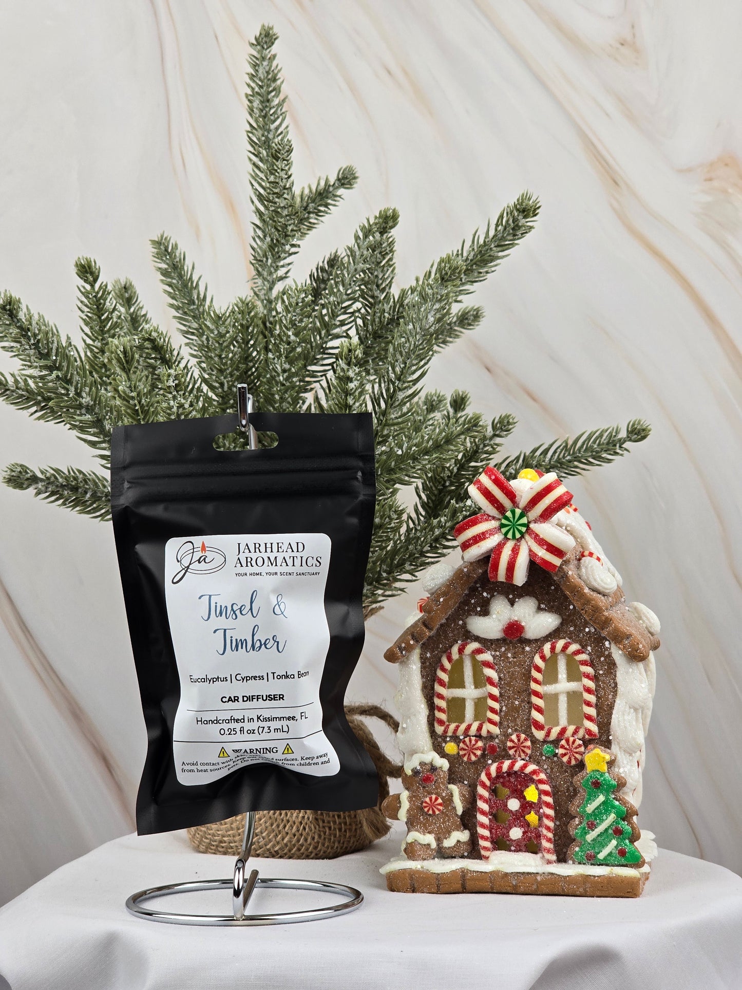 A Tinsel & Timber car diffuser from Jarhead Aromatics, in black packaging, sits on a stand next to a candy-decorated gingerbread house, releasing a festive essential oil aroma. A small Christmas tree is set against a marble-textured backdrop in the background.