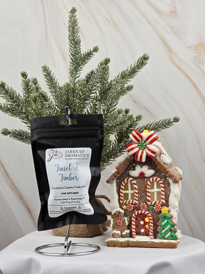A Tinsel & Timber car diffuser from Jarhead Aromatics, in black packaging, sits on a stand next to a candy-decorated gingerbread house, releasing a festive essential oil aroma. A small Christmas tree is set against a marble-textured backdrop in the background.