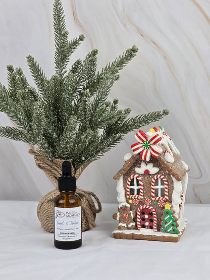 A small evergreen tree wrapped in burlap sits beside a gingerbread house with candy decorations, while a light marble background highlights the holiday scent. In front, theres a bottle of Jarhead Aromatics Tinsel & Timber - Car Diffuser Refill, enhancing the festive aroma.