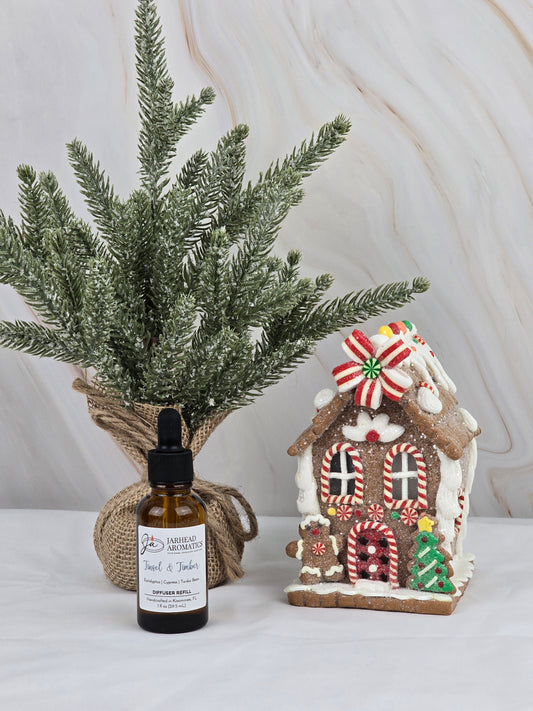 A small evergreen tree wrapped in burlap sits beside a gingerbread house with candy decorations, while a light marble background highlights the holiday scent. In front, theres a bottle of Jarhead Aromatics Tinsel & Timber - Car Diffuser Refill, enhancing the festive aroma.