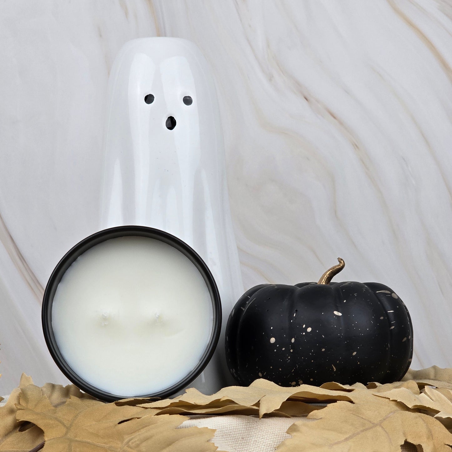 A white ghost figurine, a black pumpkin, and Jarhead Aromatics Zombie Queen Coconut Wax Blend Candle in a lit black tin are arranged with autumn leaves on a beige surface against a light marbled backdrop.