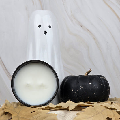 A white ghost figurine, a black pumpkin, and Jarhead Aromatics Zombie Queen Coconut Wax Blend Candle in a lit black tin are arranged with autumn leaves on a beige surface against a light marbled backdrop.