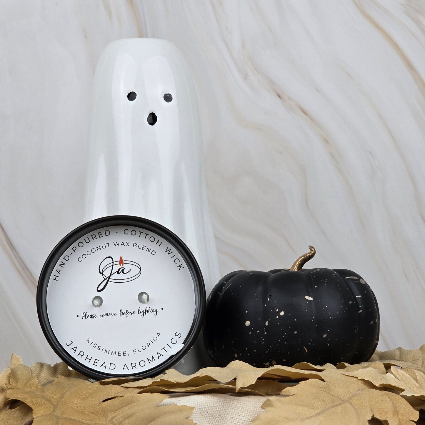 A white ceramic ghost, a black pumpkin, and the Zombie Queen - Coconut Wax Blend Candle by Jarhead Aromatics are displayed on beige fabric with brown leaves, set against a marble background.