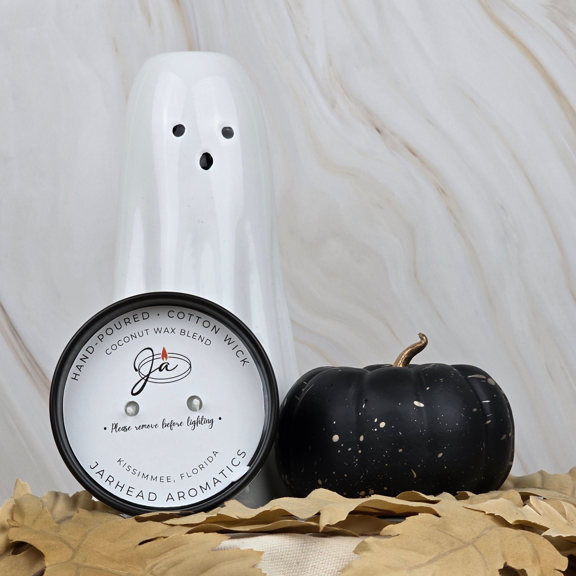 A white ceramic ghost, a black pumpkin, and the Zombie Queen - Coconut Wax Blend Candle by Jarhead Aromatics are displayed on beige fabric with brown leaves, set against a marble background.